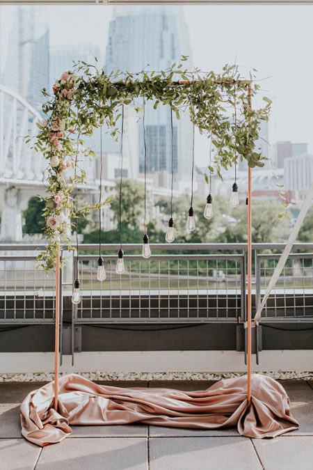 Rectangular copper ceremony arch decorated with greenery and Edison bulbs