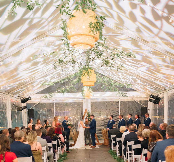 Custom leaf-print gobo shining on tent's ceiling with bamboo pendants and greenery for wedding ceremony
