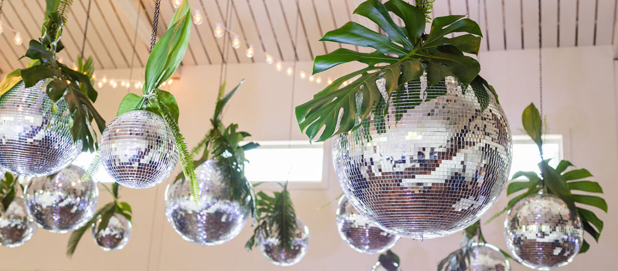 A myriad of disco balls hanging individually from the white ceiling of an event venue, each hung with large tropical leaves and fern branches